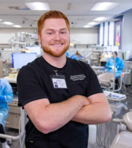 Student poses in dental clinic with several dental chair stations behind him.