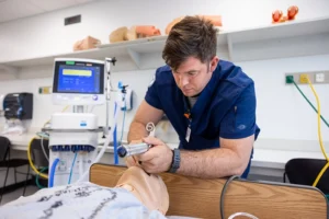 Student wearing blue scrubs stands over a simulation mannequin practicing respiratory therapy techniques