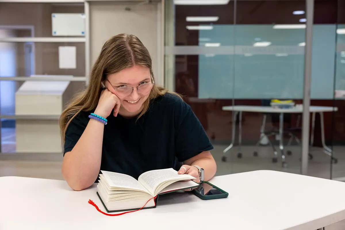 Student smiling while reading a book at a study table in the library.