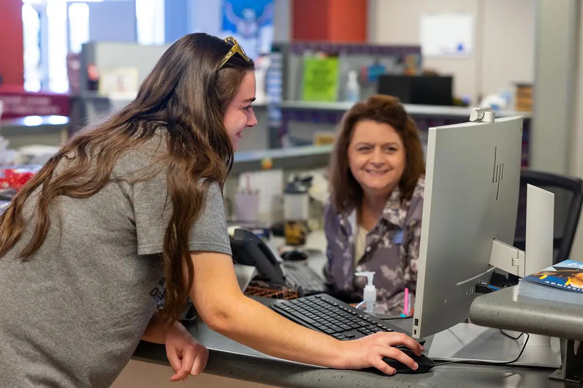 Student receiving assistance at a service desk with a staff member smiling nearby.