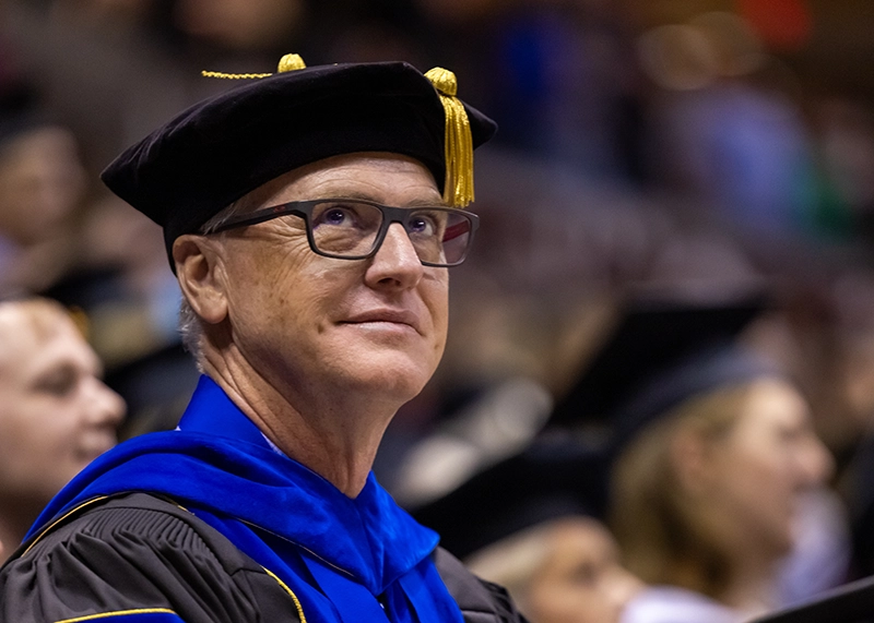 Dr. Gavin O'Connor, wearing a mortarboard, looks on at a commencement ceremony.