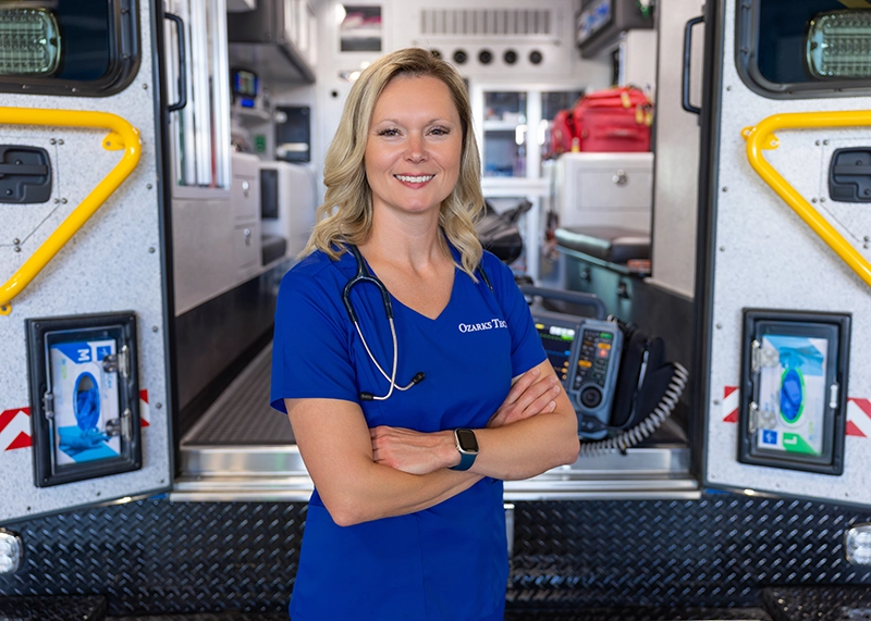 Nurse wearing scrubs stands in between the back doors of an ambulance.