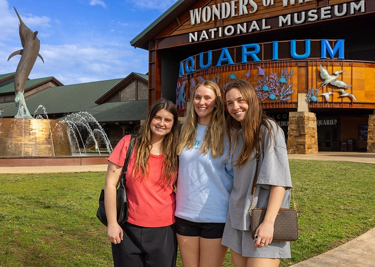 A trio of smiling young women in casual wear stand in front of the Wonders of Wildlife aquarium.