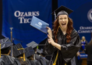 College graduate wearing a cap and gown celebrates holding up a diploma.