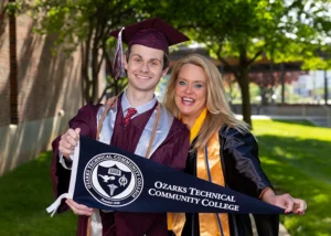 Man wearing Missouri State graduation gown stands with his mother, wearing an Ozarks Tech graduation gown. Both hold an Ozarks Tech pennant.