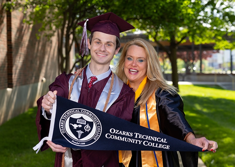 Man wearing Missouri State graduation gown stands with his mother, wearing an Ozarks Tech graduation gown. Both hold an Ozarks Tech pennant.