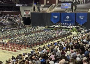 College graduates dressed in cap and gown are seated in an arena during a commencement ceremony.