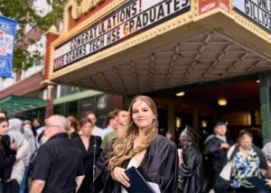 Young woman wearing a graduation gown and holding a diploma cover stands in front of a crowd at the Gillioz Theatre