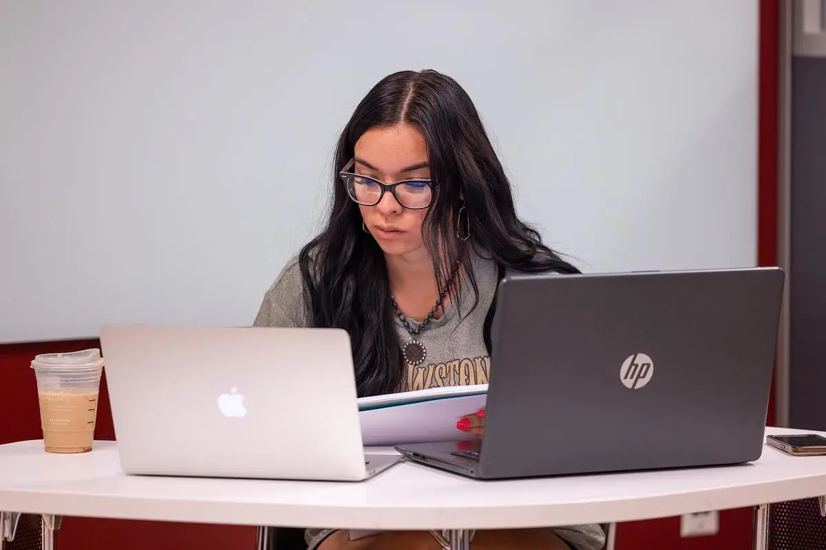 A girl sits a table in front of two laptop computers. 