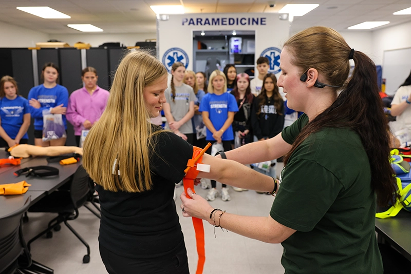 Paramedicine instructor demonstrates tourniquent application to student during hands-on training session with group of observers in classroom.