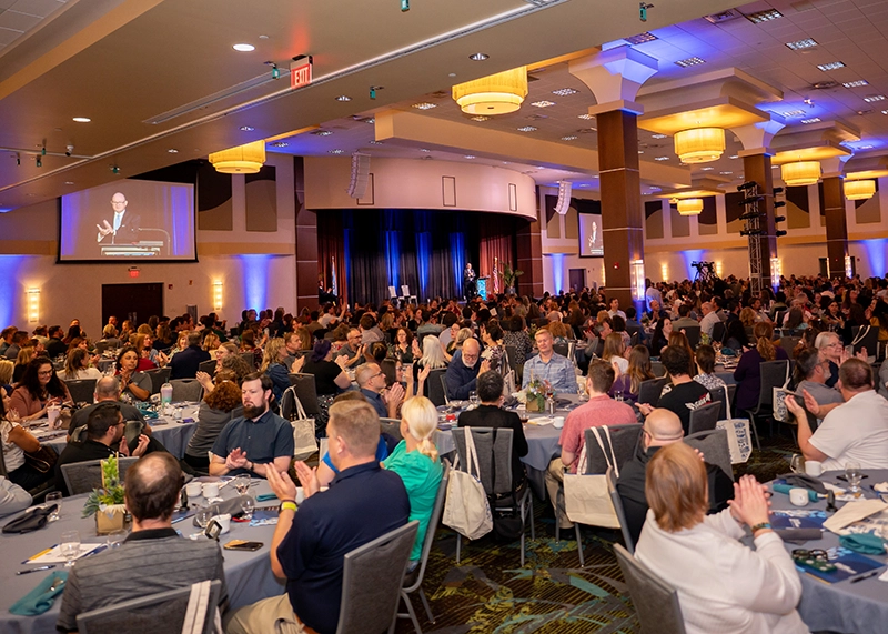 Large banquet hall filled with attendees seated at round tables during an Ozarks Tech event, with stage and blue lighting visible in background.