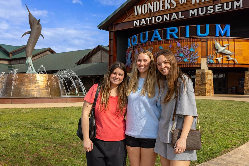 Three students pose in front of the Wonders of Wildlife aquarium building with a marlin fountain statue visible to the left.